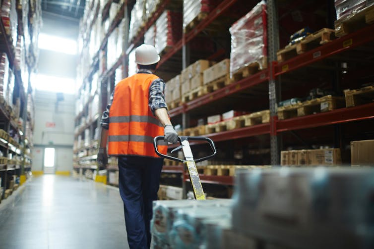 A worker in an orange vest pulls a trolley through a warehouse