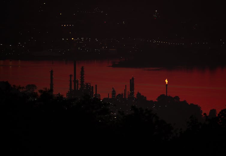 an oil refinery with a flare stack near a river with a city scape in the background at sunset.