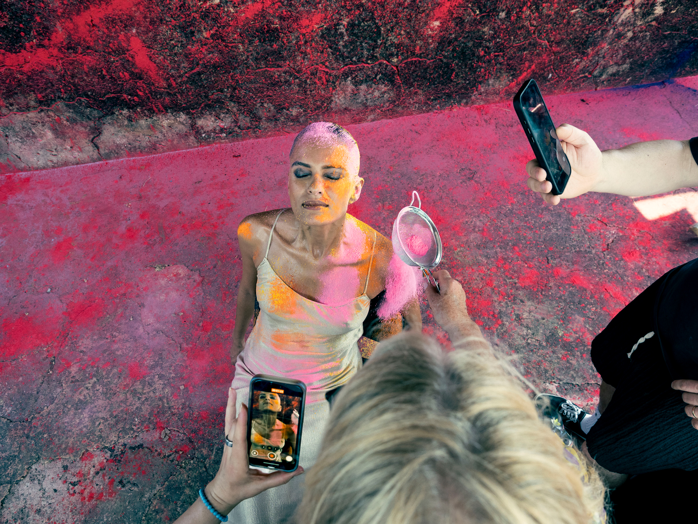 A woman sitting on the ground, around her holi color, phone and a light meter point towards her from surrounded photo assistants