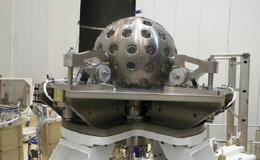 Image of a spherical silver object with regularly spaced features on its surface. It's he'd in a large room by a complicated support structure.