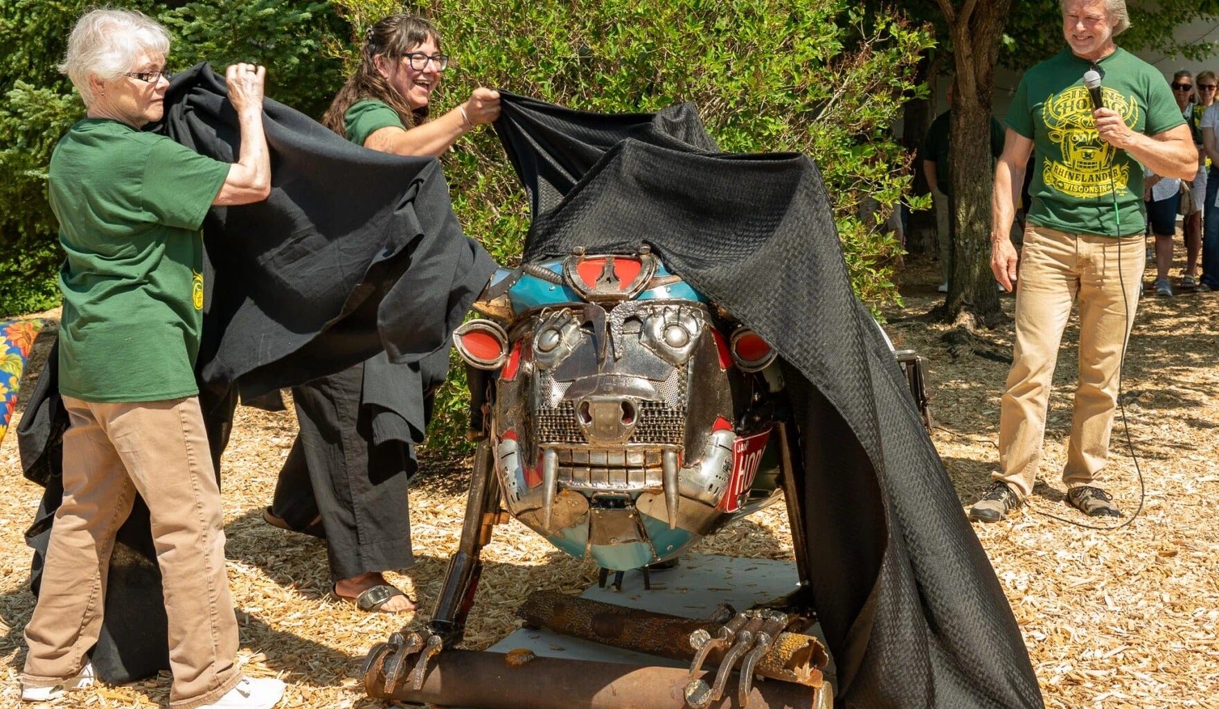 Three adults unveil a metal animal sculpture outdoors, pulling back a black cloth. Trees and wood chips are visible around them.