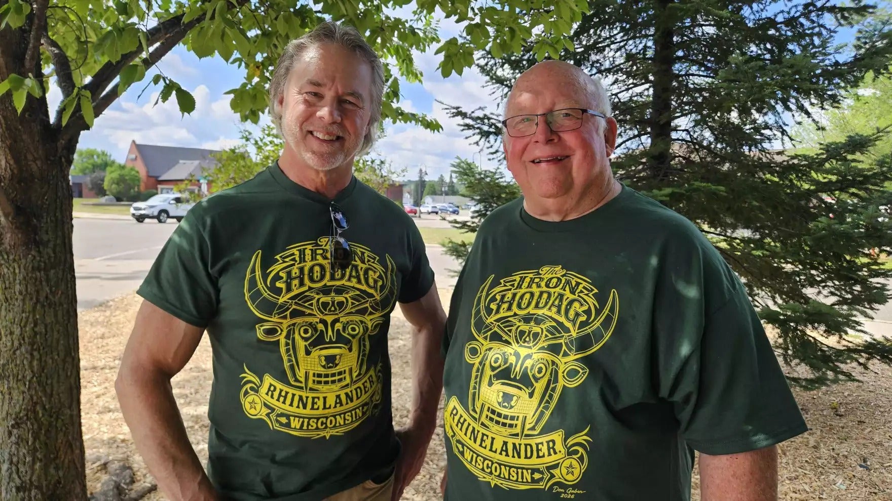 Two men stand outdoors under a tree, both wearing matching green “Rhinelander, Wisconsin” t-shirts with a yellow design.