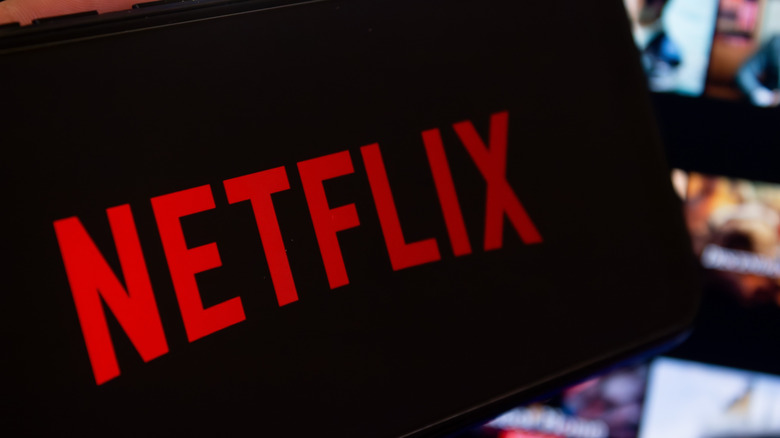 Photo of a smartphone displaying the Netflix logo. A larger screen showing media thumbnails is blurred in the background.