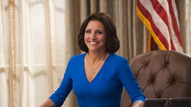 Julia Louis-Dreyfus as Selina Meyer smiling broadly while seated behind a large wooden desk, wearing a blue dress and a gold necklace in a scene from 