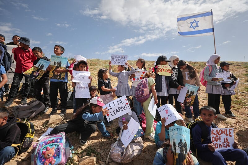 Children hold signs while standing on landscape with Israeli flag in the background