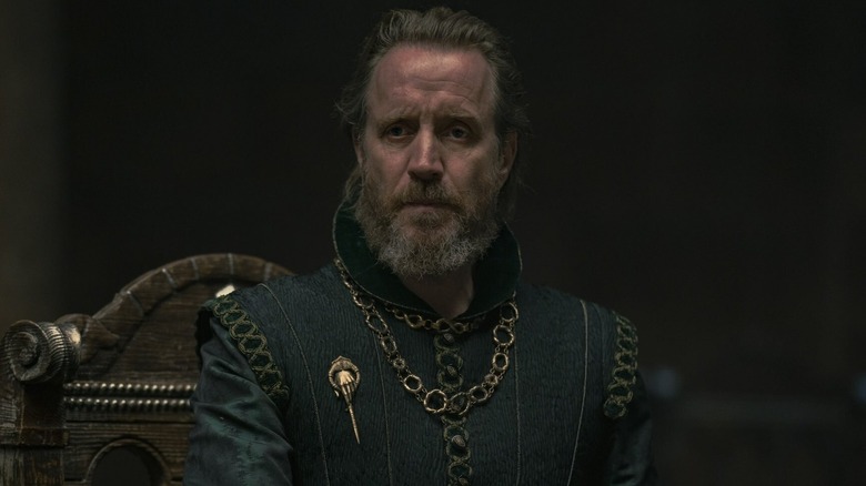 Rhys Ifans as Otto Hightower sitting and grimacing in House of the Dragon