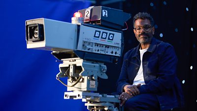 Romesh Ranganathan sits on a camera rig, operating an old BBC camera. He's pictured against a blue screen.