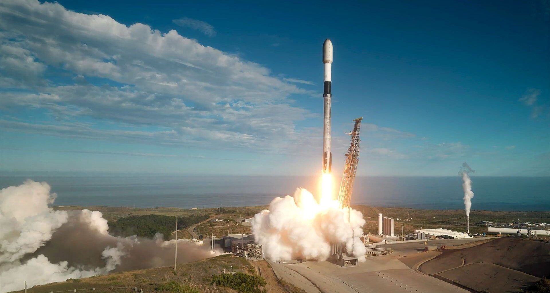 A white, black and gray-ish (soot-covered) rocket lifts off into a mostly-blue sky from a sea-side launch pad.