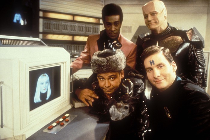 Hattie Hayridge as Holly, Craig Charles as Dave Lister, Danny John-Jules as The Cat, Robert Llewellyn as Kryten and Chris Barrie as Arnold Rimmer in Red Dwarf Dave Lister, The Cat, Kryten and Arnold Rimmer in Red Dwarf, posing next to a screen with Holly on it, all looking at the camera.
