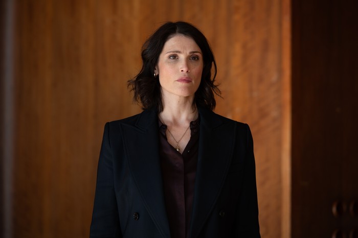 Secret Service Gemma Arterton as Kate in Secret Service, wearing a black jacket and purple shirt, stood in front of a wooden wall.