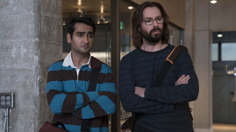 Kumail Nanjiani and Martin Starr as Dinesh and Gilfoyle standing side-by-side with arms crossed and skeptical expressions in a modern office in a scene from 