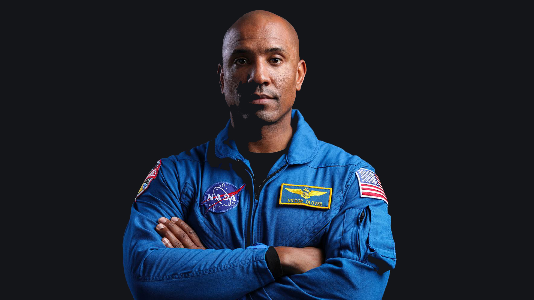 a male astronaut wearing a blue flight suit stands with his arms crossed against a dark background