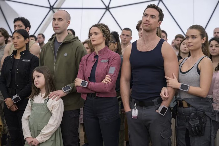 The cast of The Ark season 2, standing in a dome, looking upwards at something and looking serious, with hands in pockets and arms crossed.