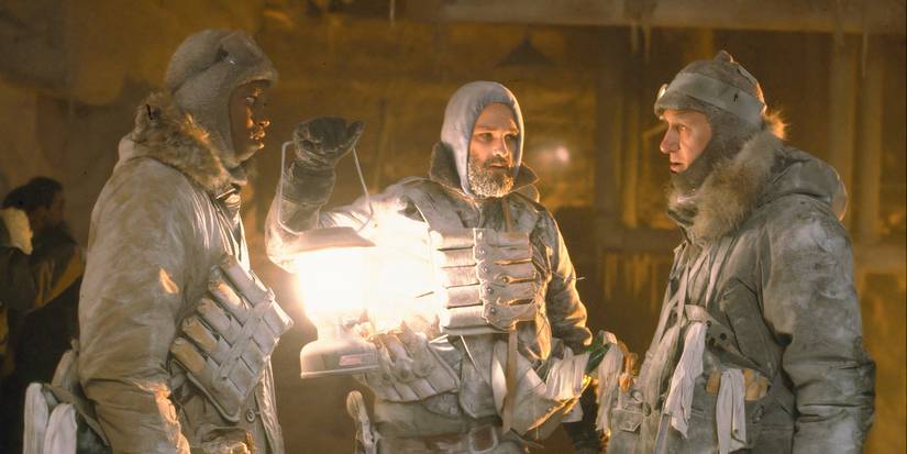 Nauls (T.K. Carter), MacReady (Kurt Russell), and Garry (Donald Moffat) standing in gear and talking while MacReady holds a lantern in The Thing