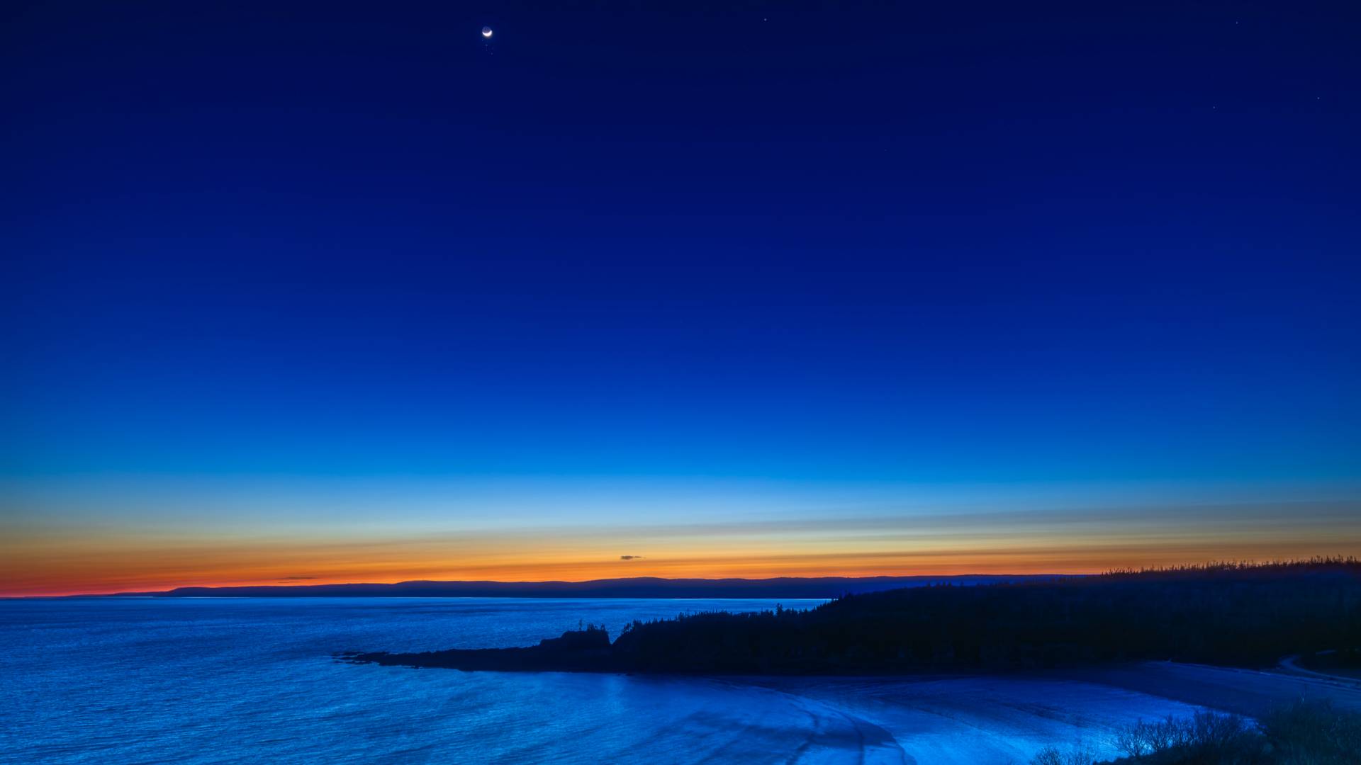 A vibrant sunset over a calm sea, with a crescent moon in the deep blue sky and an orange glow on the horizon, silhouetting dark landforms along the coastline.