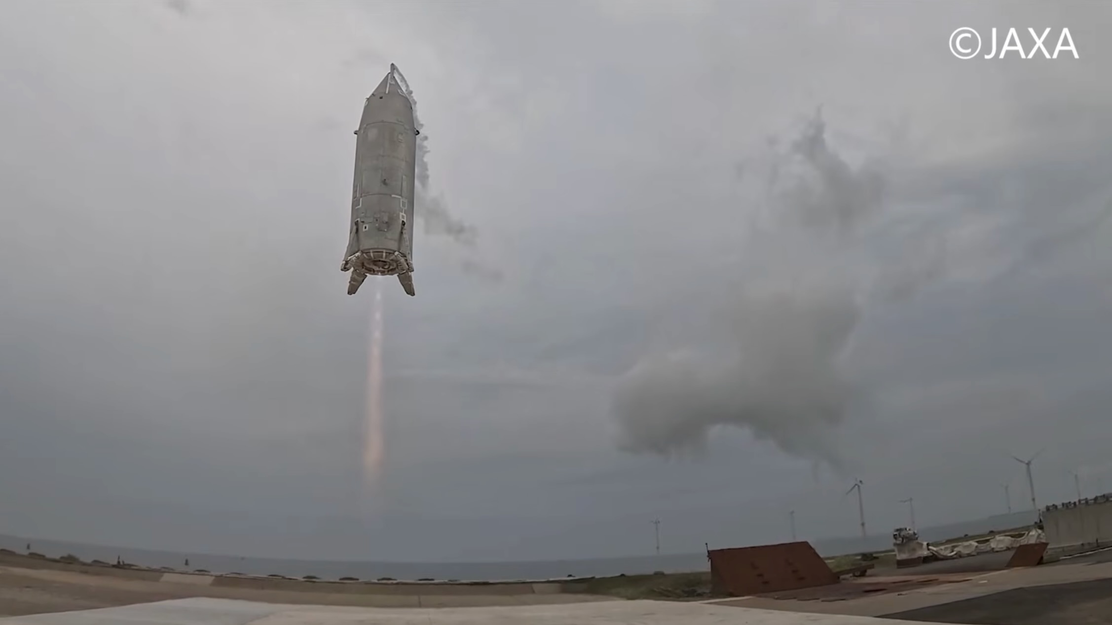 a conical silver rocket hovers about 15 feet above the ground in a cloudy sky