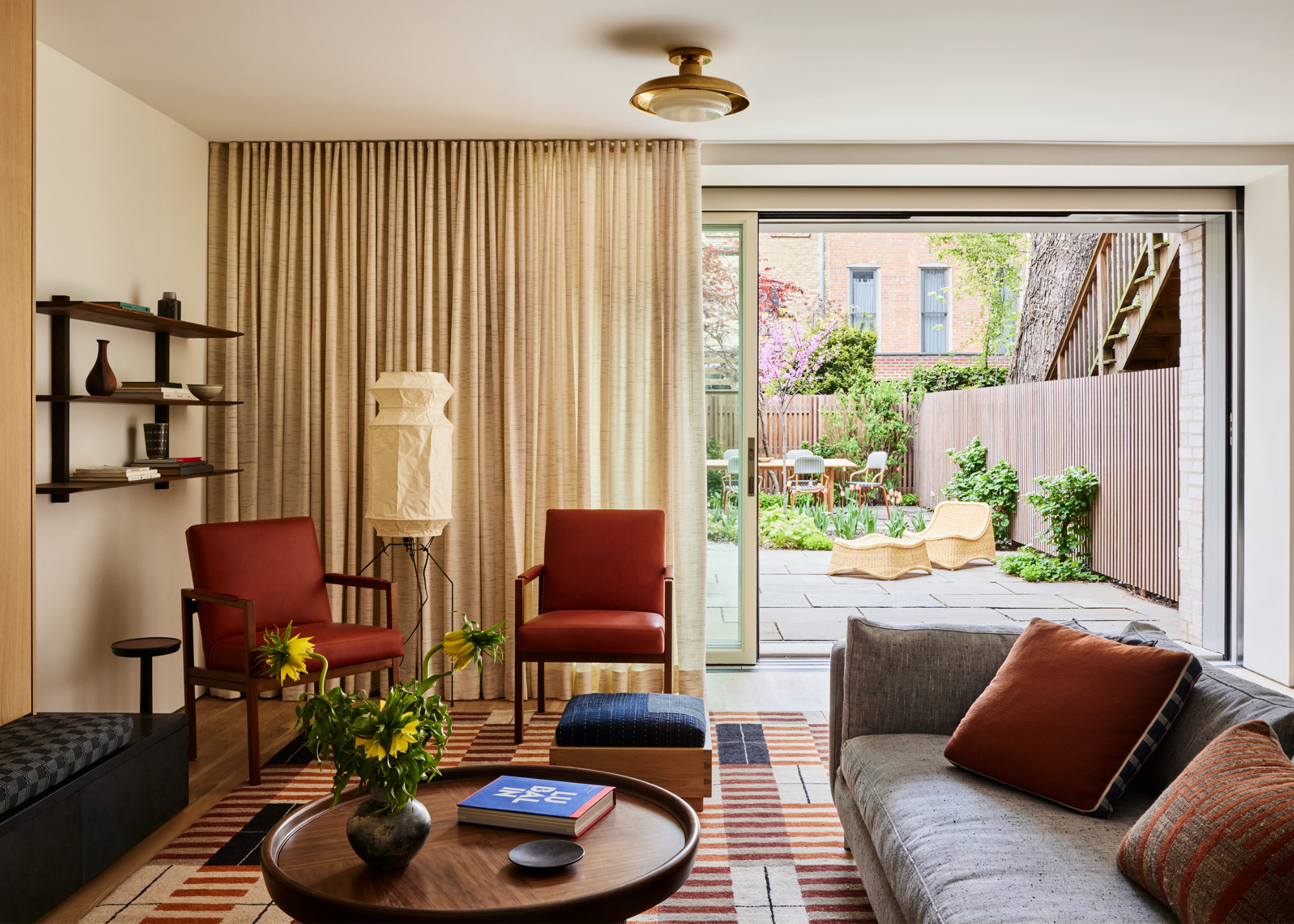 A living room with a striped area rug, a wood coffee table, a pair of rust armchairs, a gray sofa, a patterned bench, and floating shelves beside a paper floor lamp