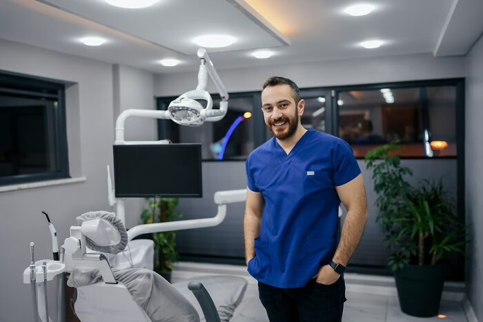 Smiling male doctor in blue scrubs standing in a modern dental office, relating to what doctors do when a patient is faking it.