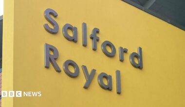 The Salford Royal Hospital sign. It is grey text emblazoned upon a yellow background.