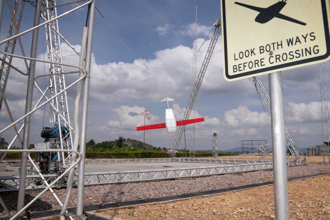 A drone, just returned from a delivery, swings in the landing mechanism in Muhanga, Rwanda. The drone is white with red wings. There is a sign that reads "look both ways before crossing" with an image of a drone.