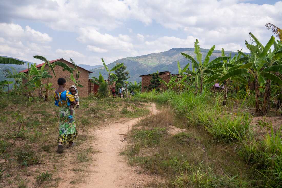 A woman holding a baby walks along a narrow dirt path that leads to Mukanoheli's village. There are several small homes with palm trees growing nearby and a mountain in the distance.