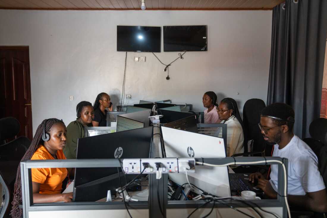 The call center in Muhanga where Zipline employees take calls and organize supplies to be sent via drone to people who need them. Six people are sitting at small desk, looking at computer monitors and wearing headsets.
