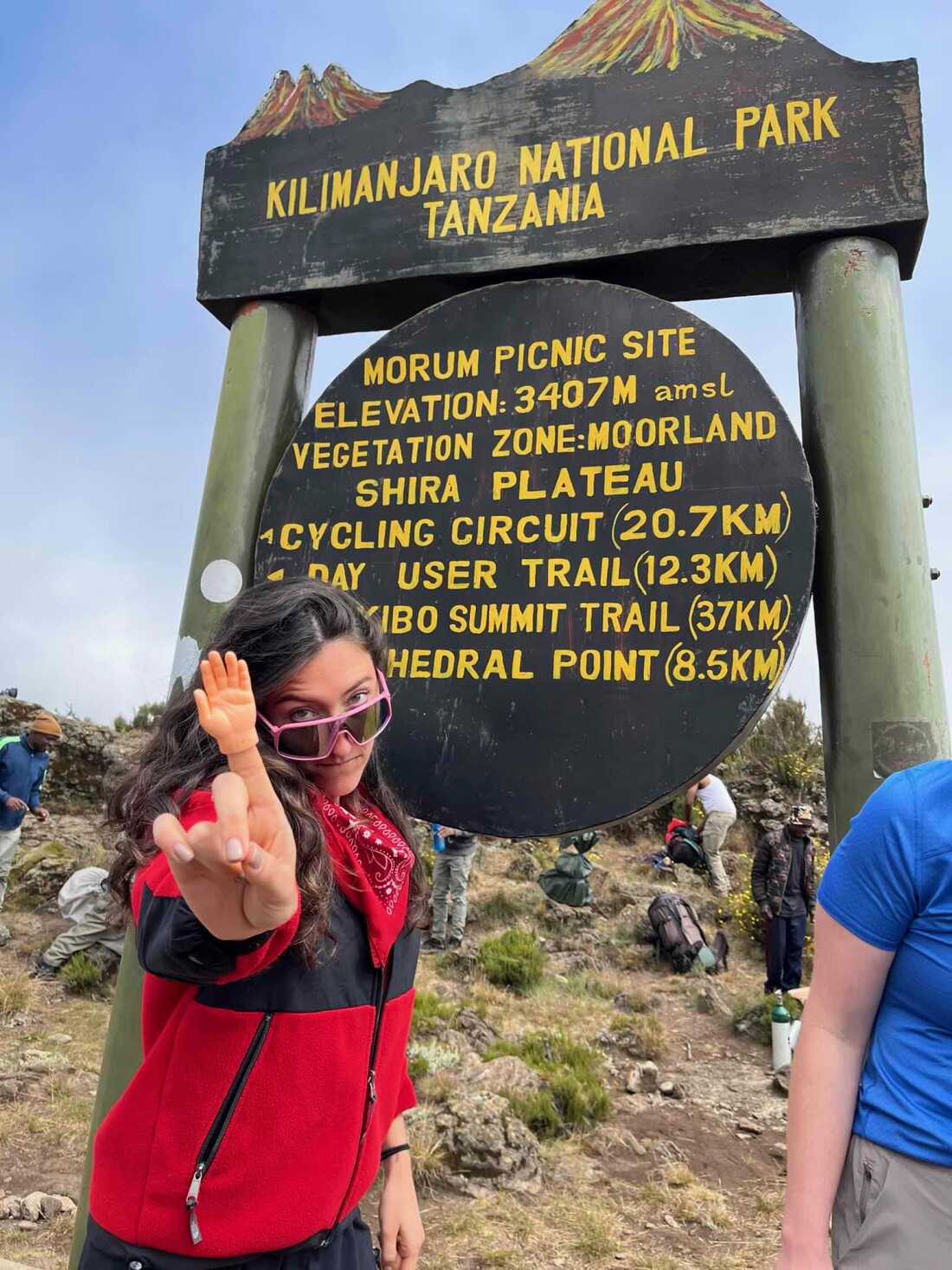A vibrant young woman is shown in front of a sign for Kilimanjaro National Park, Tanzania. She is looking over her sunglasses and gesturing at the camera with a finger puppet on her index finger.