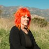 Micky Small, 53, is a screenwriter and is one of hundreds of millions of people who regularly use AI chatbots. She has bright orange, shoulder length hair and is seated with a background of mountains and brush behind her.