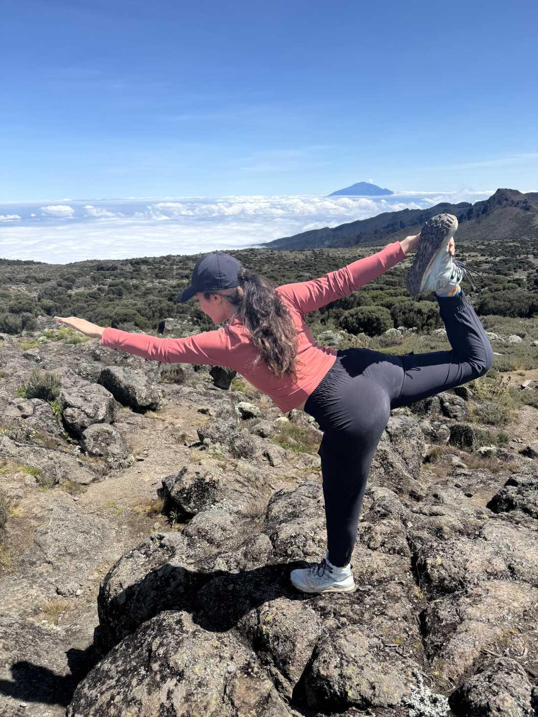 Sophie Rottenberg is shown with a rugged mountain vista behind her. She is balanced on one leg in yoga's "dancer" pose.