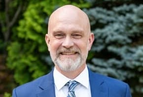 A man in a blue suit stands smiling at the camera.