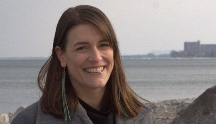 A woman in a grey coat stands in front of a lake smiling.