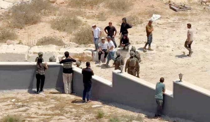 Palestinians use their phones as Israeli settlers and military personnel stand outside a Palestinian home in Qusra. (REUTERS)