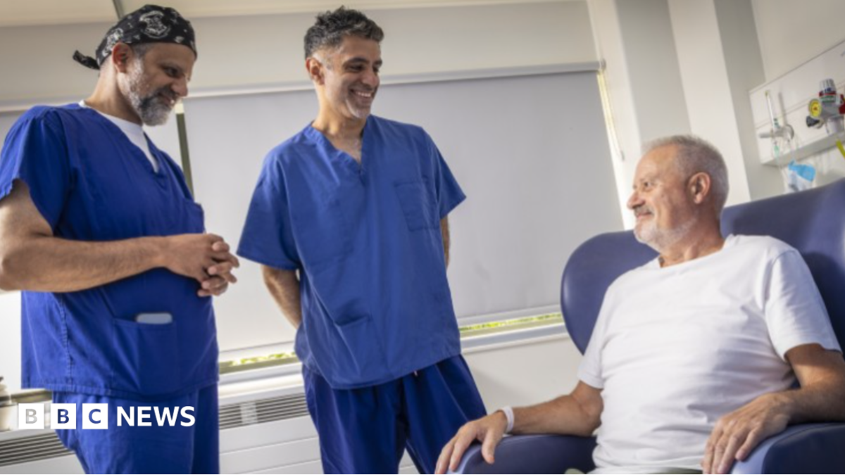 Two men dressed in blue scrubs are on the left and in the middle and standing up. They are looking towards Hall, who has a beard and grey hair and is sitting on a blue chair on the right.