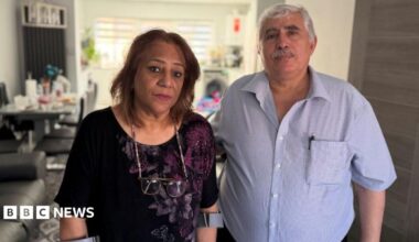 Farah and Nihad Fathi. She has shoulder length brown hair and is wearing a black top with a purple floral pattern. He ha short grey hair and a moustache with a blue checked shirt open at the collar. They are standing together in their home.