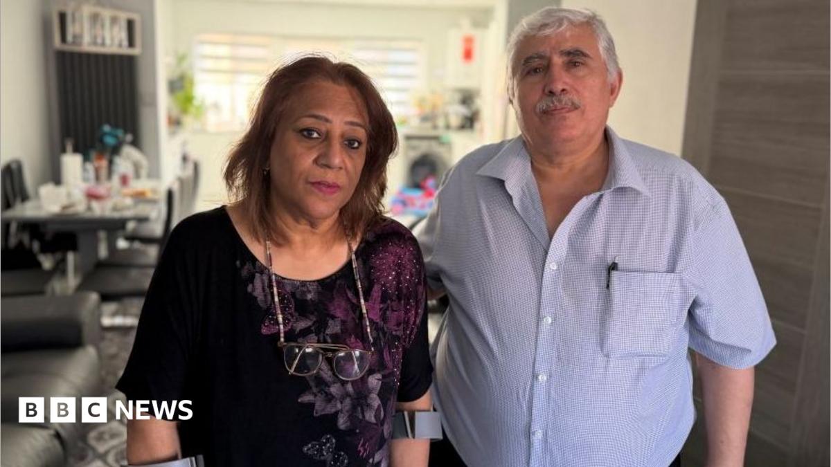 Farah and Nihad Fathi. She has shoulder length brown hair and is wearing a black top with a purple floral pattern. He ha short grey hair and a moustache with a blue checked shirt open at the collar. They are standing together in their home.