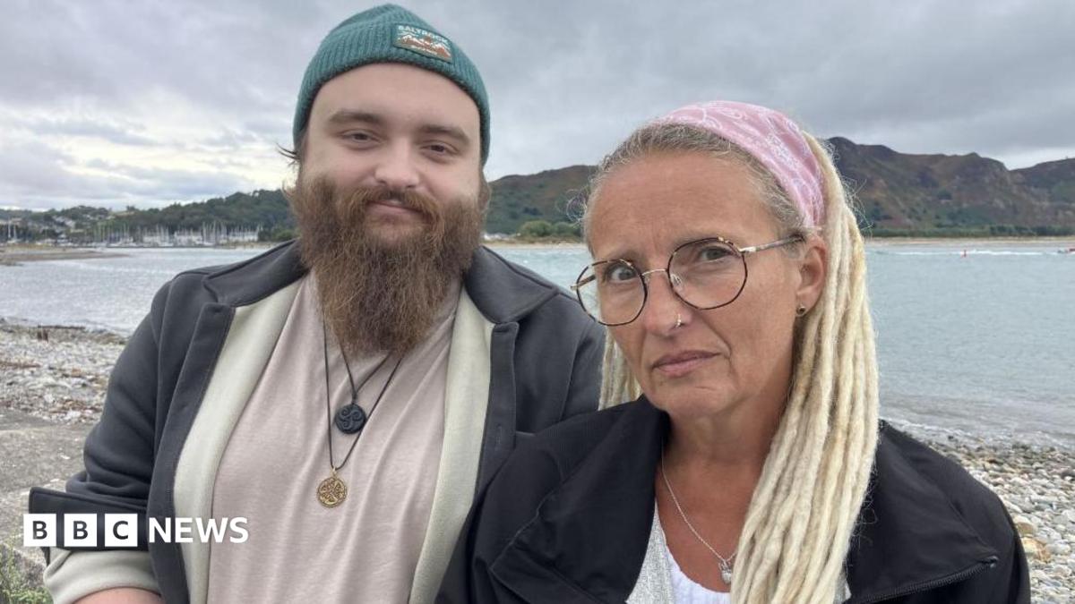 Ryan Shields and his mother Aly Shields standing together looking towards the camera. His mother is wearing a pink bandana on her blonde hair and has brownish glasses. Ryan is wearing greyish jacket and a blue beanie. He also has a long, light brown beard.