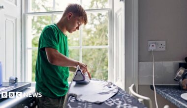 Stock photo shows a person ironing a T shirt in front of a window in their kitchen, next to a plug socket and with the washing machine behind them.
