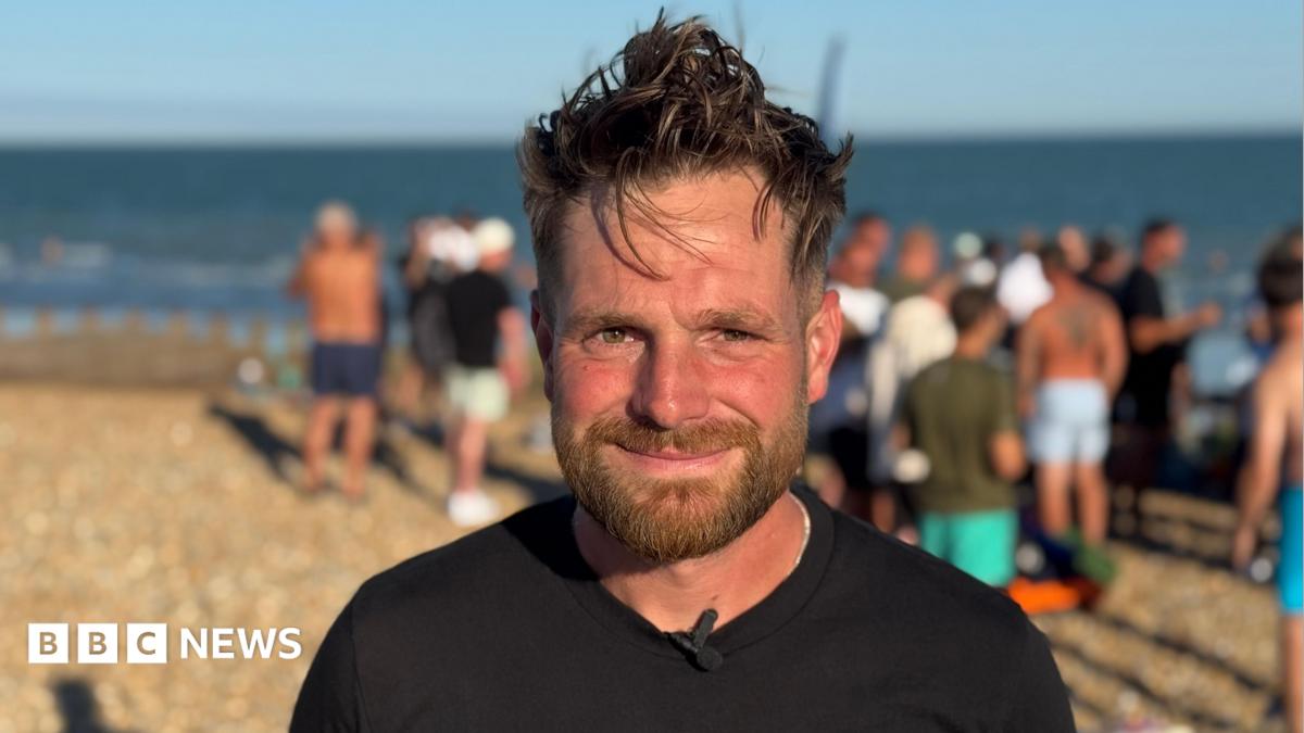 Joe smiles to the camera. He is stood on a pebbly beach with a blue sea behind him and a group gathered on the beach behind him too. It is sunny.