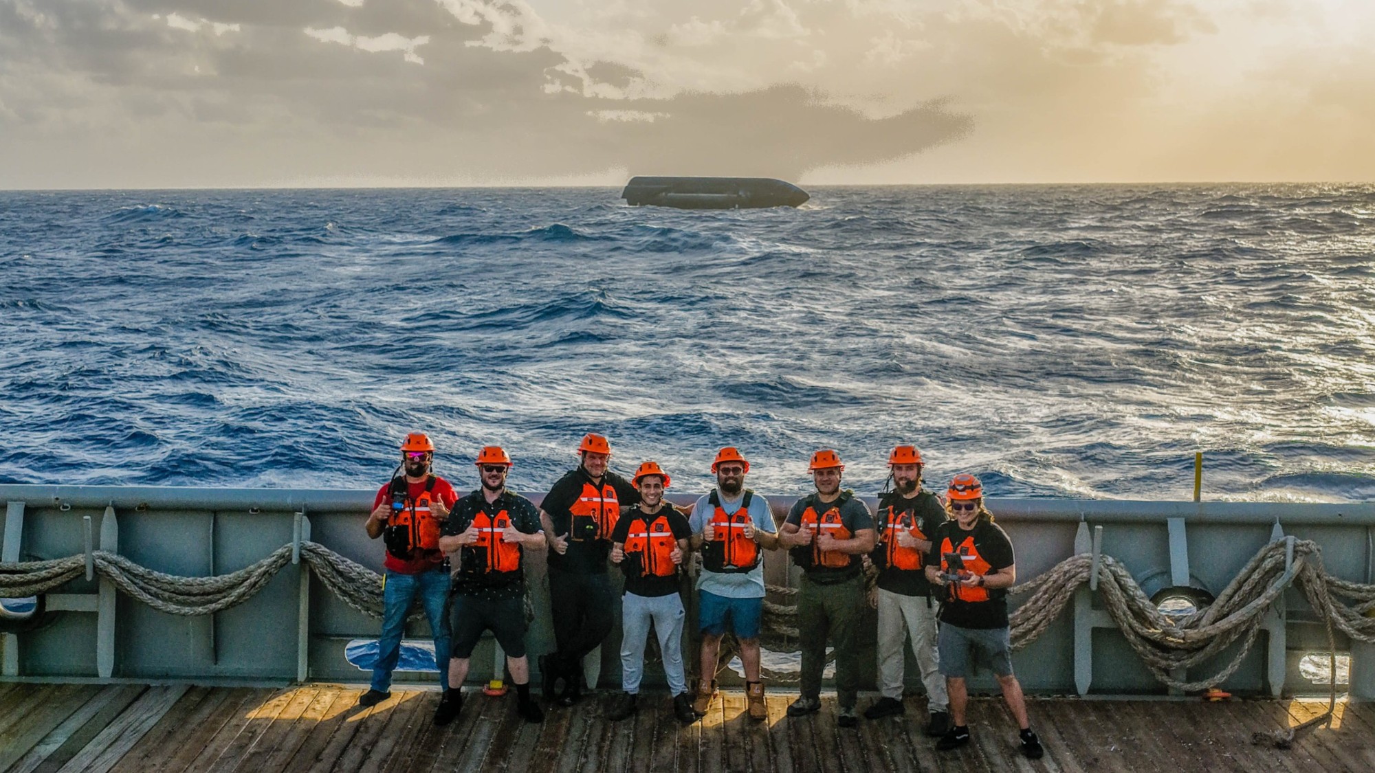 eight people stand on the deck of a ship at sea. behind them in the distance, a large black cylinder floats on top of the waves