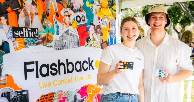 A young woman and man stand in front of a colorful collage backdrop branded "Flashback," each holding a small, rectangular camera.