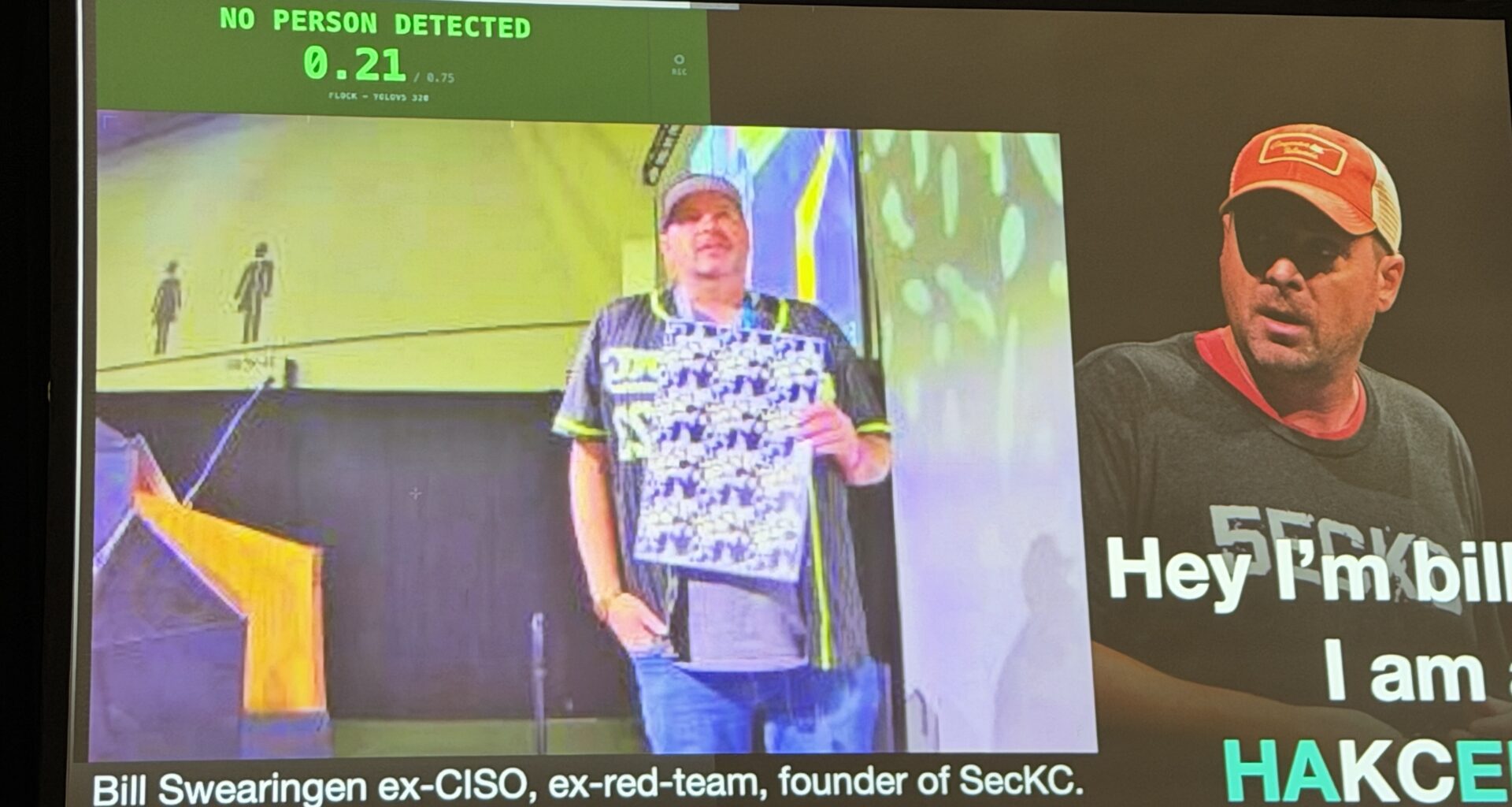 A person on a stage holds a pattern chest high in an attempt to hide his identity from AI detectors. Behind him, a screen show that the AI did not detect a person.