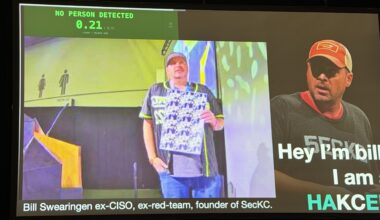 A person on a stage holds a pattern chest high in an attempt to hide his identity from AI detectors. Behind him, a screen show that the AI did not detect a person.
