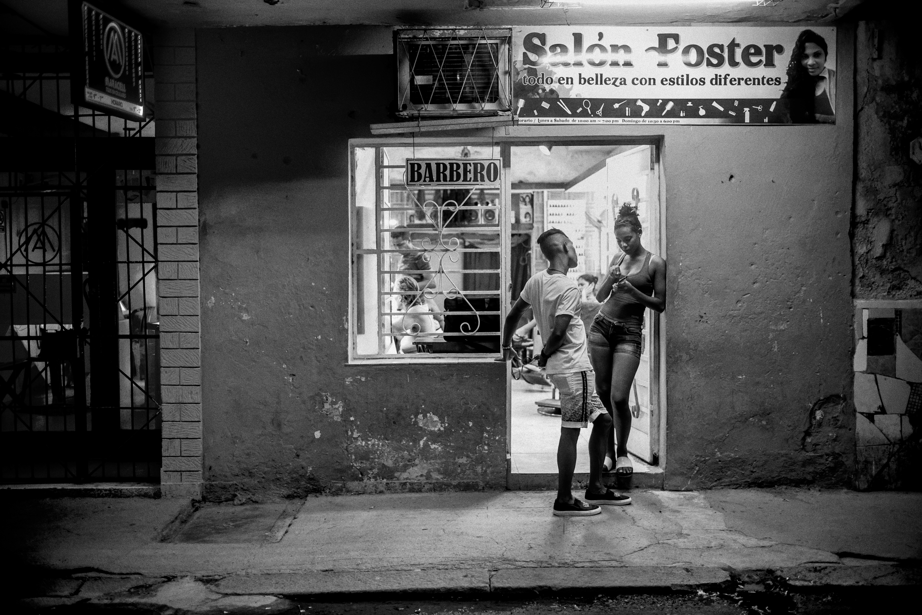 Black and white night shot of a young couple standing outside a hair salon in Havana, its illuminated sign reading "Salón Foster, todo en belleza con estilos diferentes."
