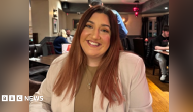 A woman looking at the camera. She has long brown hair and smiles at the camera. She is wearing a beige blazer with a brown top underneath.