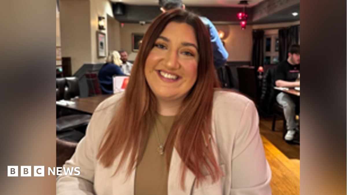 A woman looking at the camera. She has long brown hair and smiles at the camera. She is wearing a beige blazer with a brown top underneath.