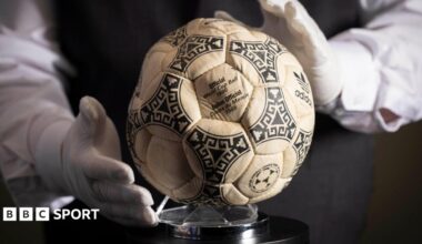 A person wearing white gloves places the football used in the 1986 World Cup quarter-final on a stand
