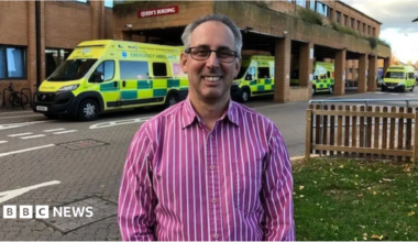 A man in a pink striped shirt stands in front of a hospital emergency department . Behind him are several ambulances.