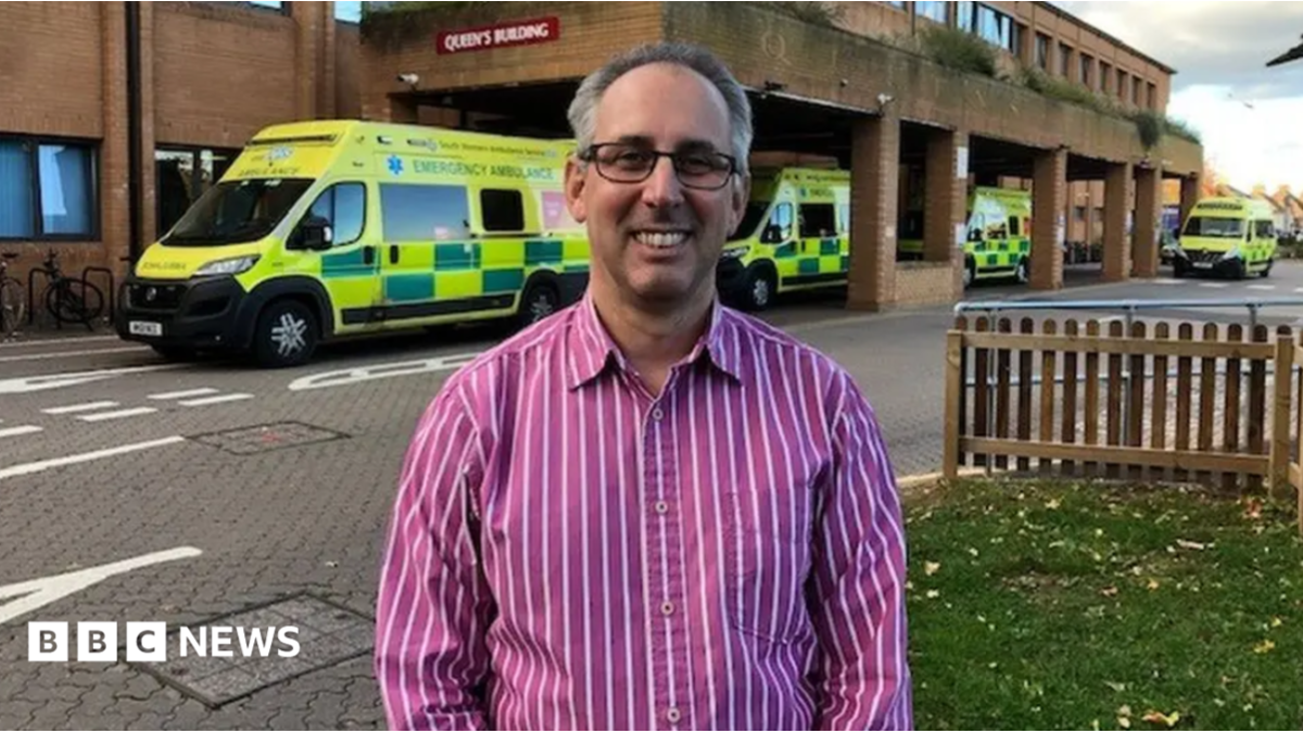 A man in a pink striped shirt stands in front of a hospital emergency department . Behind him are several ambulances.
