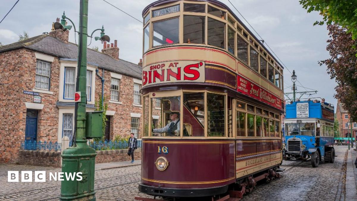 Visitors travel on a vintage electric tram in the 1900s village at Beamish Museum. There is a row of terraced houses on a cobbled street. Two double-decker public transport vehicles from different eras are in shot. The front one is a maroon and beige tram with a curved front and a large advert for Binns between the two floors. The rear one has more of the appearance of an old trolleybus or lorry, with a cab for the driver, an exposed radiator and distinct tyres. It is bright blue and open-topped.