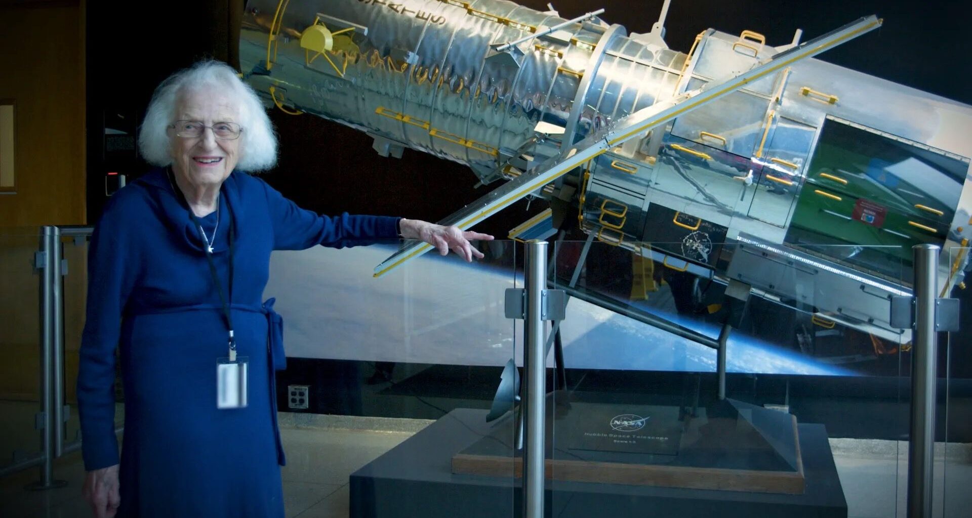 an elderly white-haired woman in a blue dress stands next to a model of a silver space telescope
